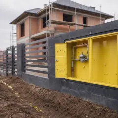 Żółta skrzynka z instalacją gazową w murze, obok budowanego domu i ogrodzenia.