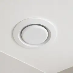Biały, okrągły anemostat wywiewny zamontowany na białym suficie.