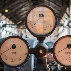 Trzy zabytkowe mierniki: VOLT, AMPERE i KILOWATT, wskazujące wartości prądu i mocy.