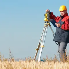 Geodeta w żółtym kasku i kurtce pracuje z niwelatorem. Zastanawiasz się, ile kosztuje geodeta?