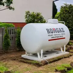 Duży, biały zbiornik na propan firmy ROMGAZ stoi na fundamencie w ogrodzie.