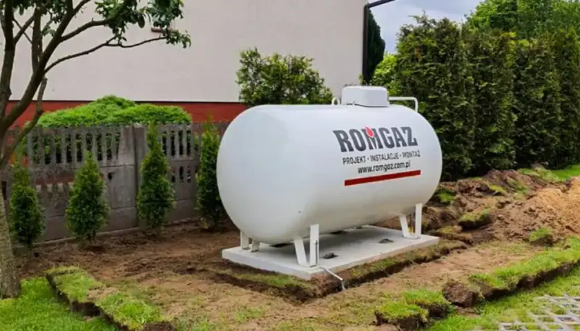 Duży, biały zbiornik na propan firmy ROMGAZ stoi na fundamencie w ogrodzie.