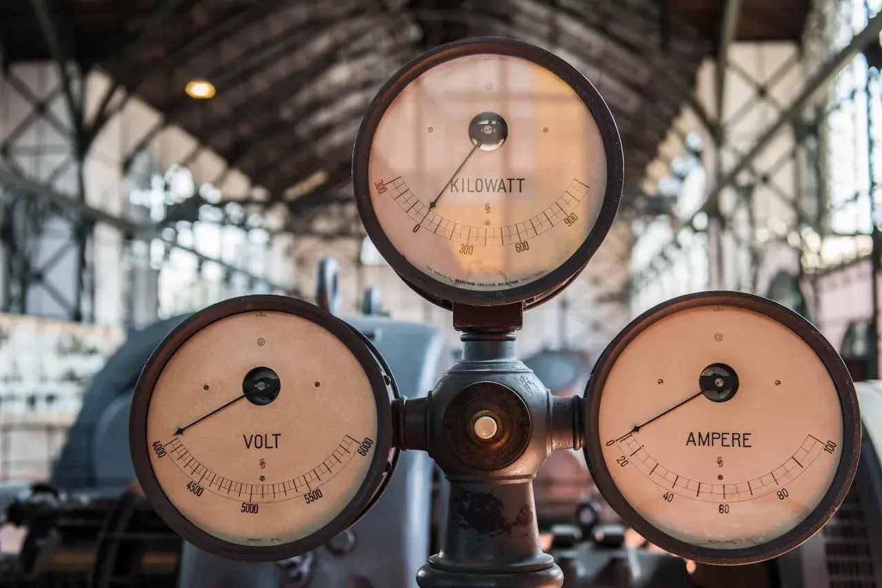 Trzy zabytkowe mierniki: VOLT, AMPERE i KILOWATT, wskazujące wartości prądu i mocy.