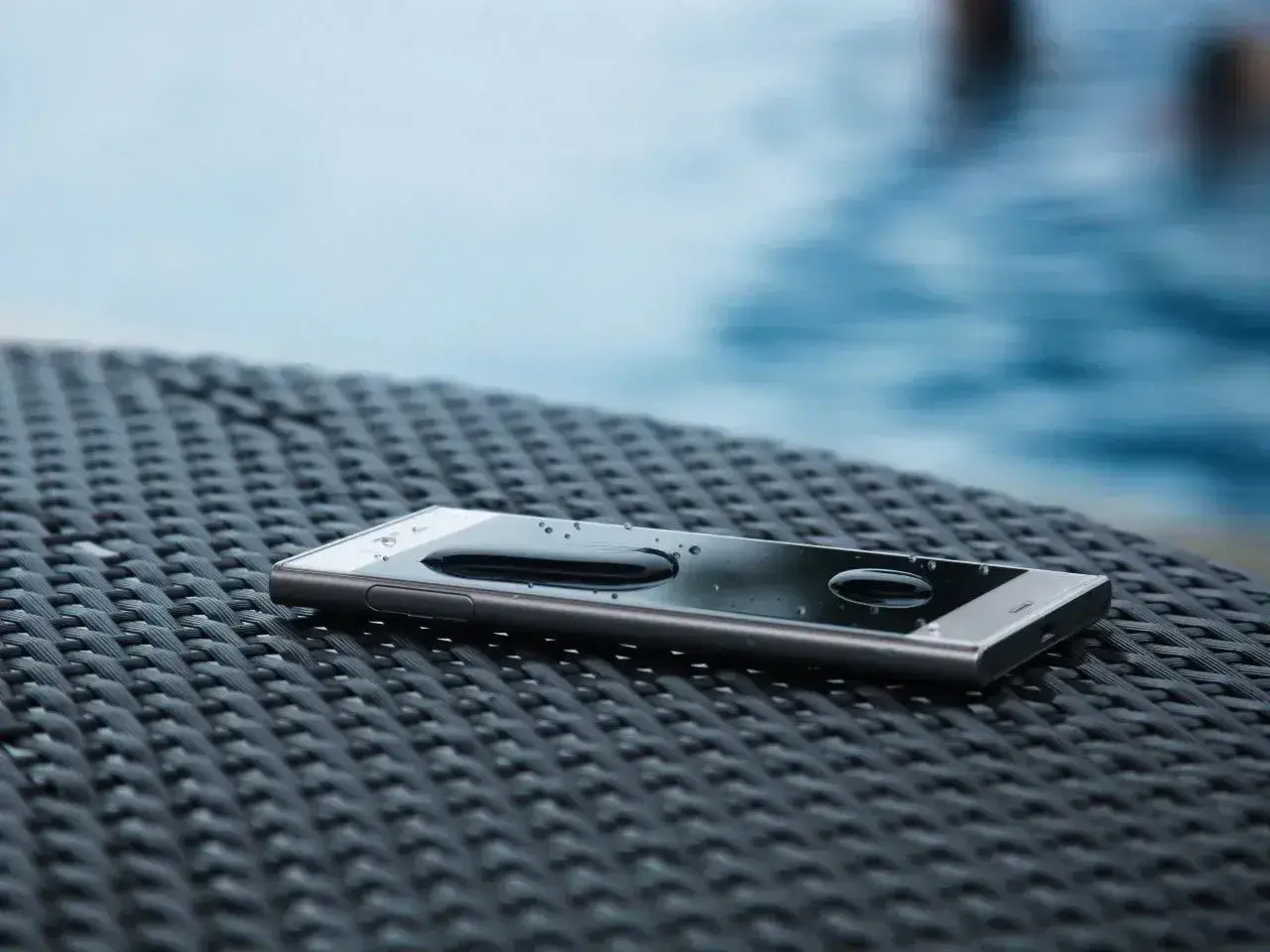 Telefon z kroplami wody na ekranie, leżący na plecionej powierzchni obok basenu. To dowód, że ip68 co oznacza – pełną ochronę przed wodą.