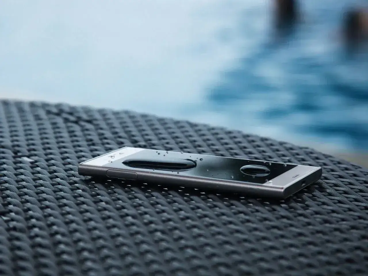 Smartfon z certyfikatem IP44 leży na stoliku przy basenie, krople wody na ekranie świadczą o jego wodoodporności.